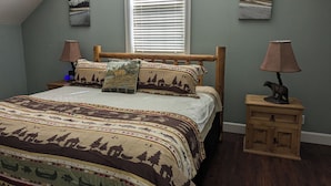 A bedroom with a bed, two bedside tables, a window with blinds, and two wall-mounted paintings.
