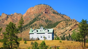 A two-story house with a green roof, surrounded by a grassy field and pine trees, with a rocky mountain in the background.