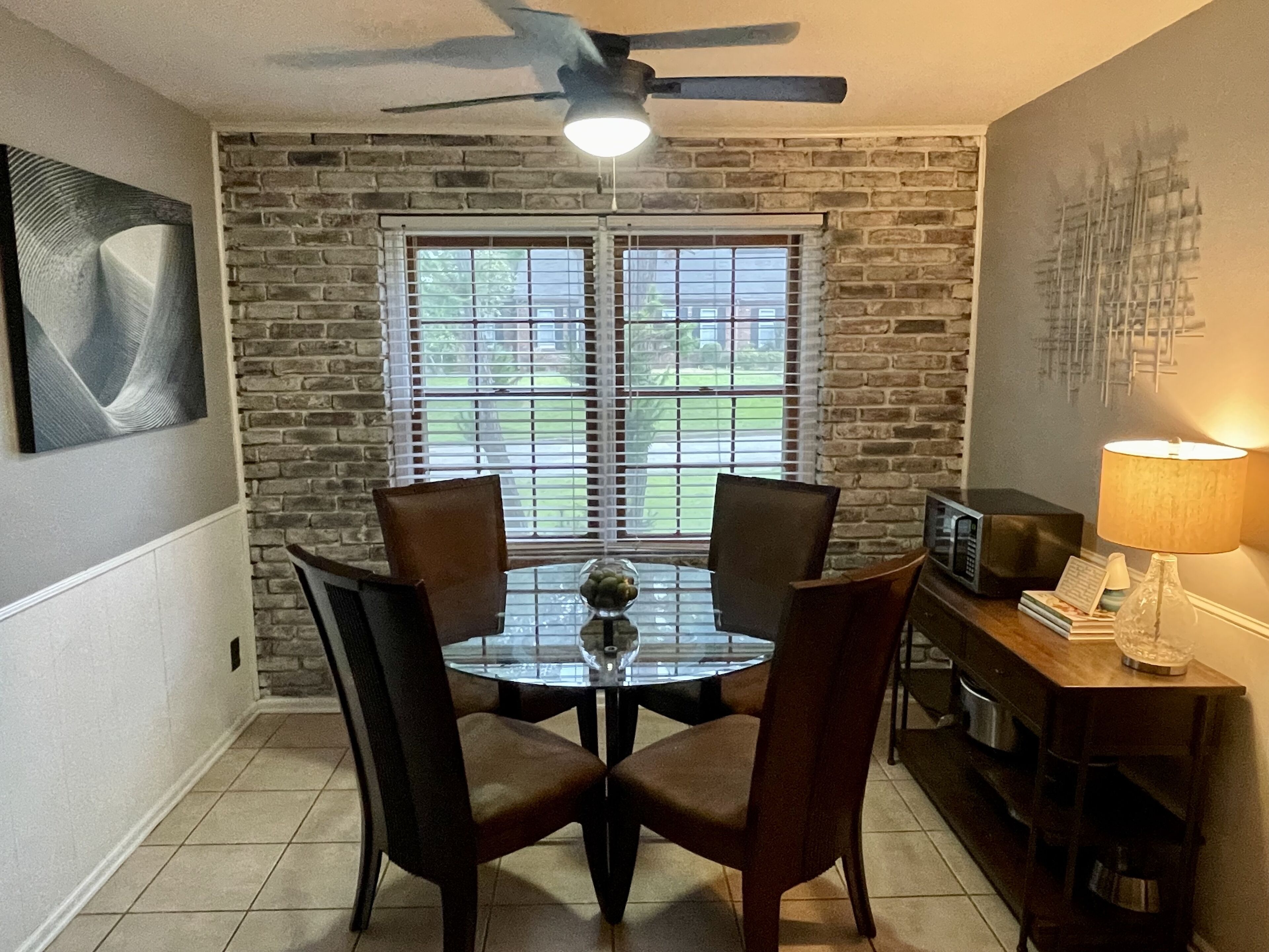 A dining area with a brick wall, two windows with blinds, a ceiling fan, a table with chairs, and a television on a stand.