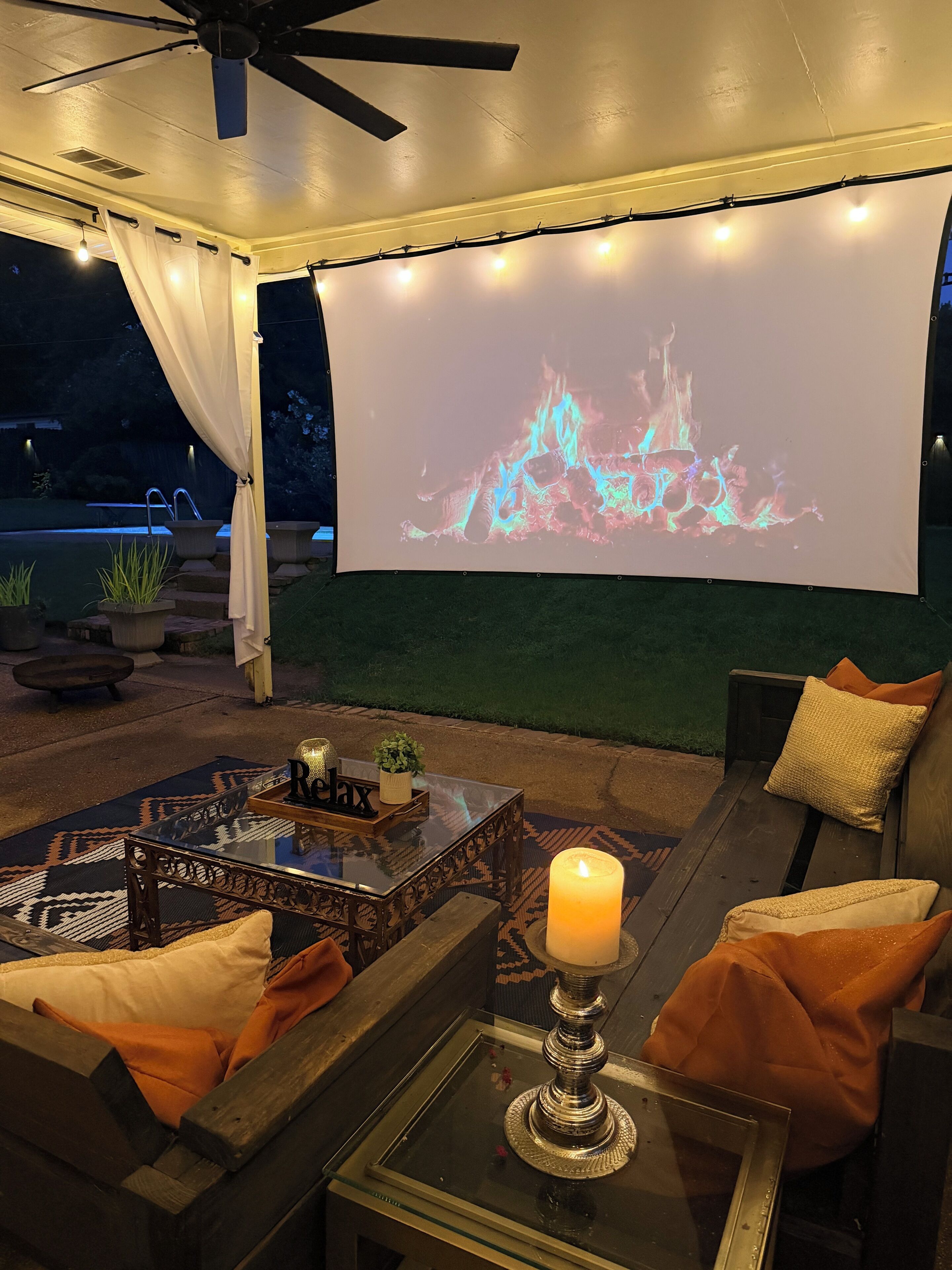 An outdoor patio with a ceiling fan, a projection screen displaying a fire, and a seating area with cushions and a table.