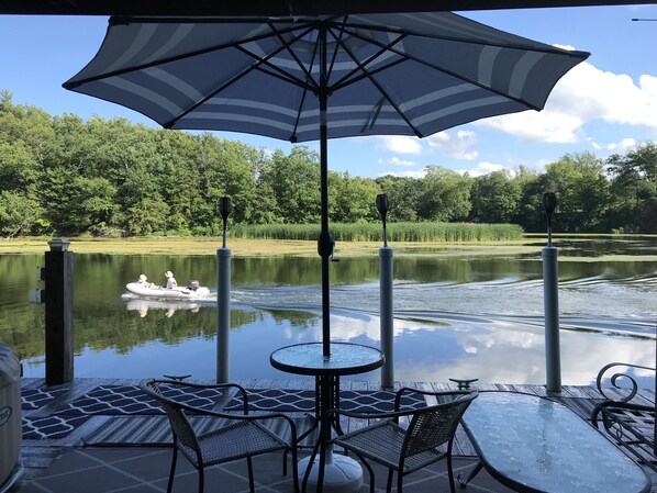 Ein Außenbereich zum Sitzen mit Tisch und Stühlen, einem großen Sonnenschirm und Blick auf einen See mit einem Boot.
