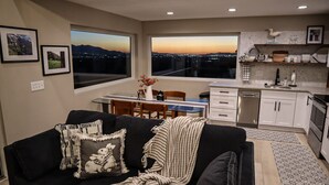 En moderne stue med en sort sofa, et spisebord og et køkken med hvide skabe samt et stort vindue med udsigt over en by ved solnedgang.