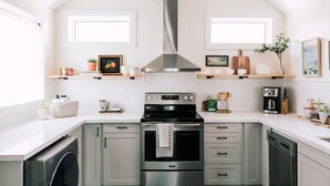 Een moderne keuken met een roestvrijstalen afzuigkap, een ingebouwde oven en een vaatwasser. Boven de aanrecht zijn twee ramen en de muren zijn wit geschilderd.