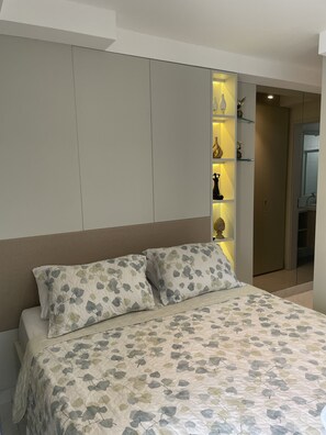 A bedroom with a bed, two pillows, a bedside table, and a shelf with decorative items. - Pé na areia do Caribessa - Branco Haus (João Pessoa)