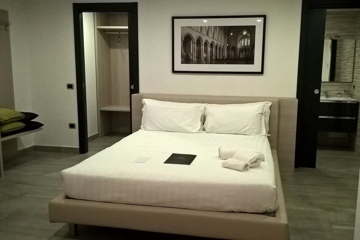 A neatly made bed with pillows and a folded towel, a framed picture above it, and a bathroom visible through an open door.
