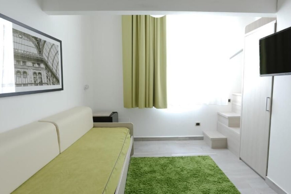 A modern room with a white sofa, a green rug, a window with curtains, a flat-screen TV, and a staircase leading to an upper level.