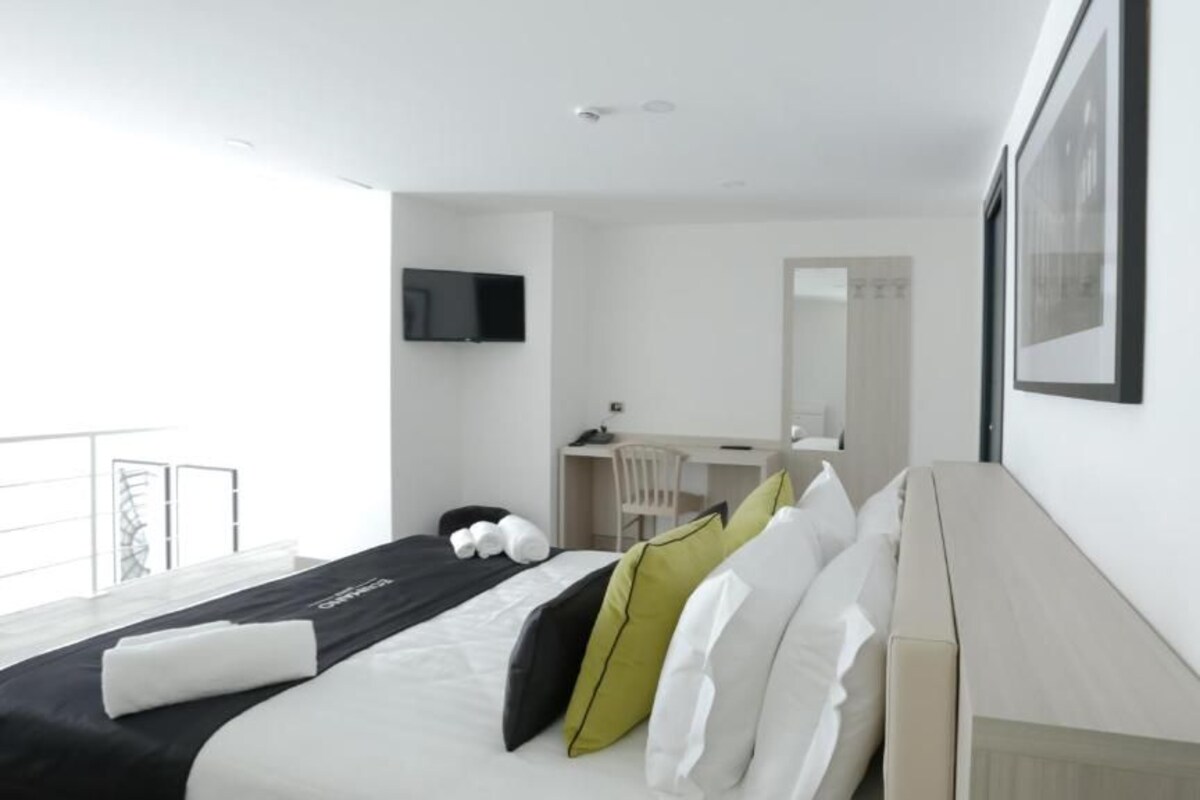 A modern hotel room with a large bed, a desk, a chair, a TV, and a balcony.
