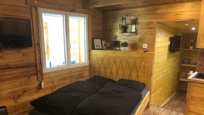 Ein Zimmer in einer Holzhütte mit einem Bett, einem Fernseher, einem Fenster und einer Küchenzeile.