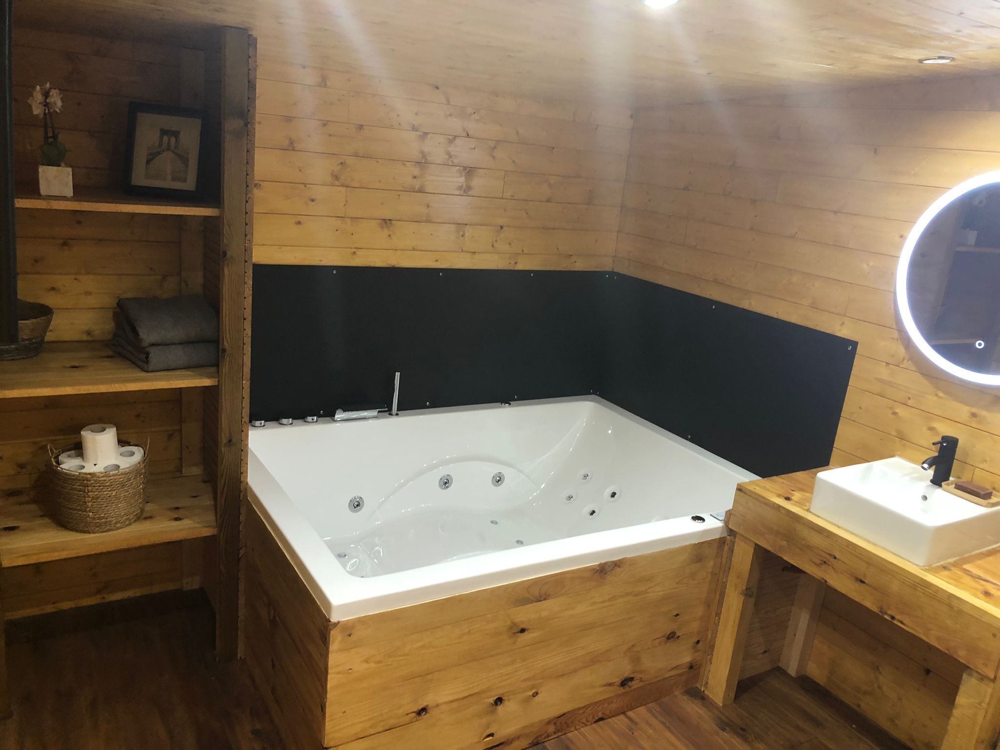 Een houten badkamer met een groot bad, een klein wastafeltje en planken met toiletartikelen.