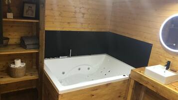 Een houten badkamer met een groot bad, een klein wastafeltje en planken met toiletartikelen.