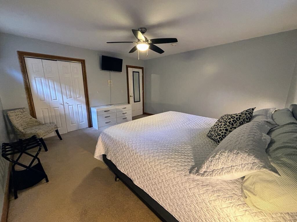 A bedroom with a bed, a ceiling fan, a TV on the wall, and a door.
