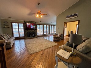 A spacious living room with hardwood floors, a stone fireplace, a ceiling fan, and a television. - Family-Fun Escape w/ Pool, Game Room, Firepit + Baby Gear (Corbin)