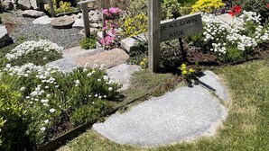 A wooden signpost with the text "USE AT OWN RISK" in a garden area with rocks, flowers, and a view of a lake with boats. - Historic Oceanfront Home with private dock and boat house. Walkable to town. (Southwest Harbor)