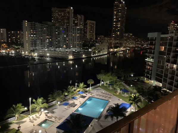 Enjoy a serene waterfront view from the comfort of our pool area, surrounded by illuminated buildings and a tranquil body of water.