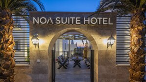 Indgangen til Noa Suite Hotel med en buet portåbning, udendørs siddeområde og to vægmonterede lamper.