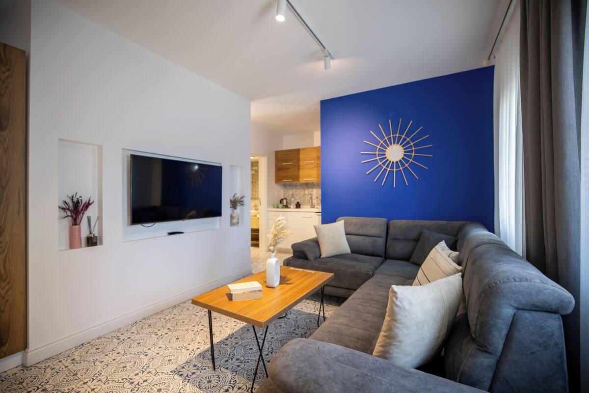 A modern living room with a grey sofa, a wooden coffee table, a flat-screen TV, and a blue accent wall with a sunburst design.