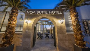 Indgangen til NOA SUITE HOTEL med en stenbuet indgang, palmer og et udendørs siddeområde.