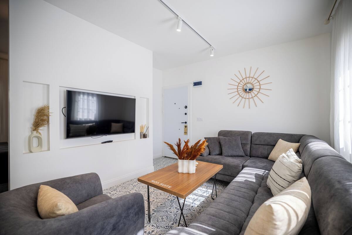 A modern living room with a grey sofa, a wooden coffee table, a flat-screen TV, and a decorative sunburst wall piece.
