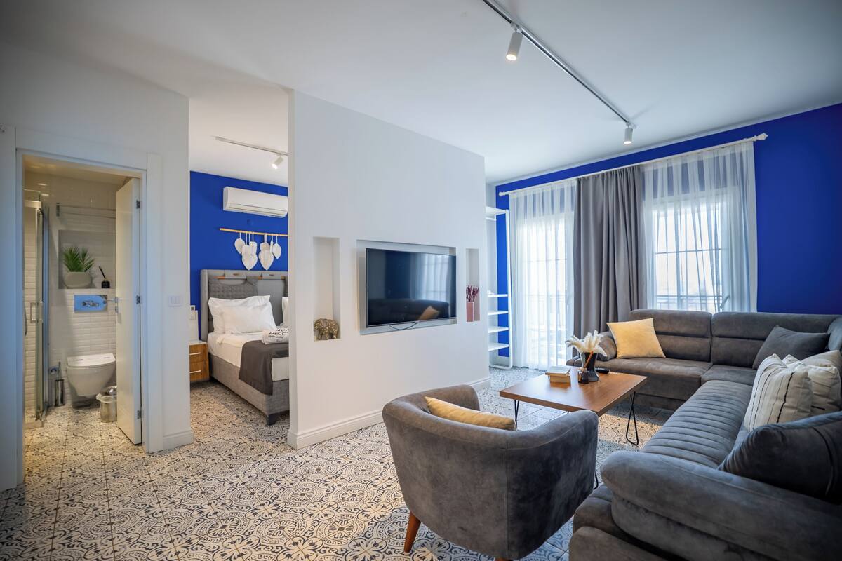 A modern living room with a grey sofa, a small armchair, a TV mounted on the wall, and a large window with curtains.