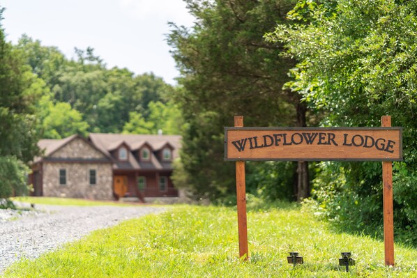 A wooden sign with the text "WILDFLOWER LODGE" in front of a stone building with a red roof. - Luxury Lodge Fire Pit, Hot Tub, & Sauna (Front Royal)