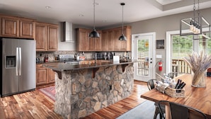 Una cocina con armarios de madera, una isla de piedra, electrodomésticos de acero inoxidable, iluminación colgante y vistas al exterior.