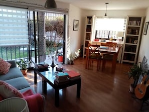 Sala de estar com sofá, mesa de centro e livros. Há uma área de jantar com mesa e cadeiras. Uma guitarra é visível no canto.