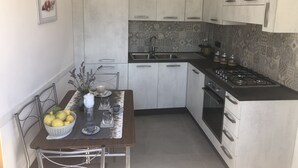 A compact kitchen with white cabinets, a black countertop, and a gas stove. There is a dining table with a basket of lemons and a vase with flowers. - Appartamento nel Cuore Della Costa Damalfi (Pietre)