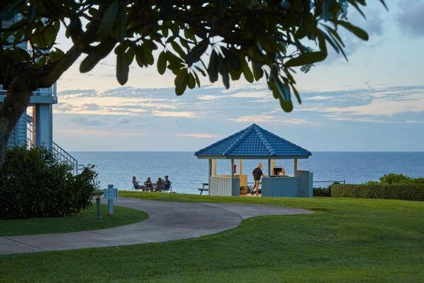 A gazebo with a blue roof overlooking a body of water, a pathway leading to it, and a building with a staircase on the left.
