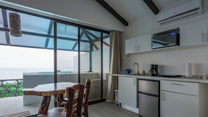 A modern kitchen with a built-in microwave, a coffee maker, and a small refrigerator. There is a dining area with a wooden table and chairs, and large windows offering a view of the ocean. - Akua Ocean View Suites (Tárcoles)
