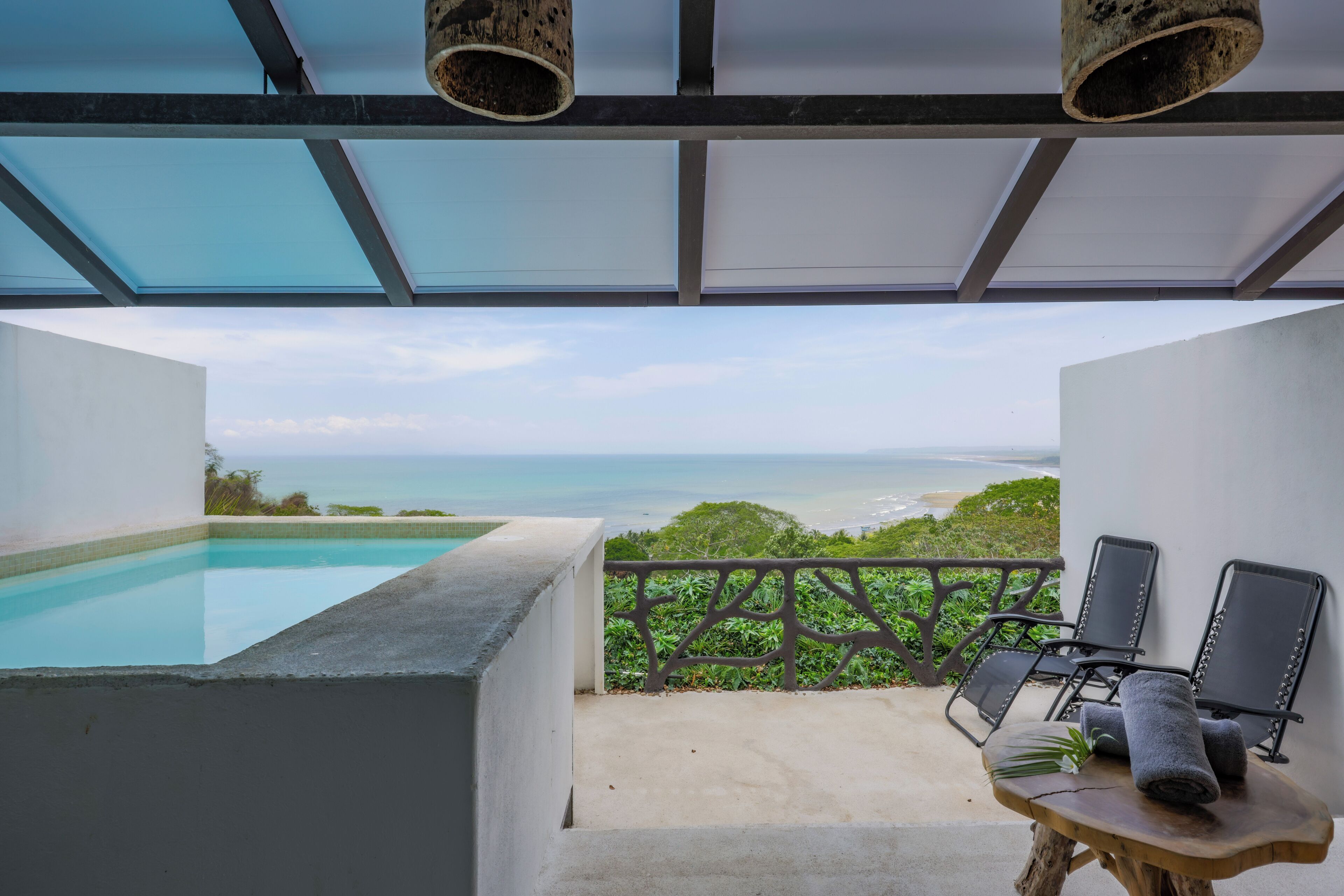 A balcony with a view of the ocean, a pool, and two lounge chairs.