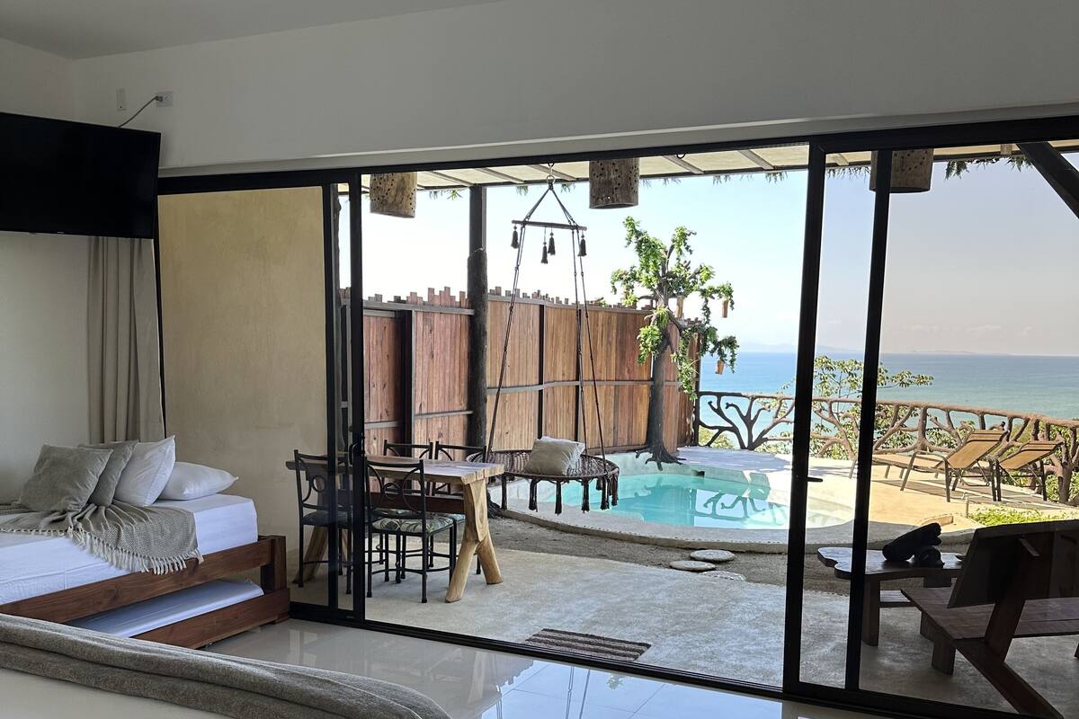 A bedroom with a large bed, a dining area with a table and chairs, and a view of a pool and beach.