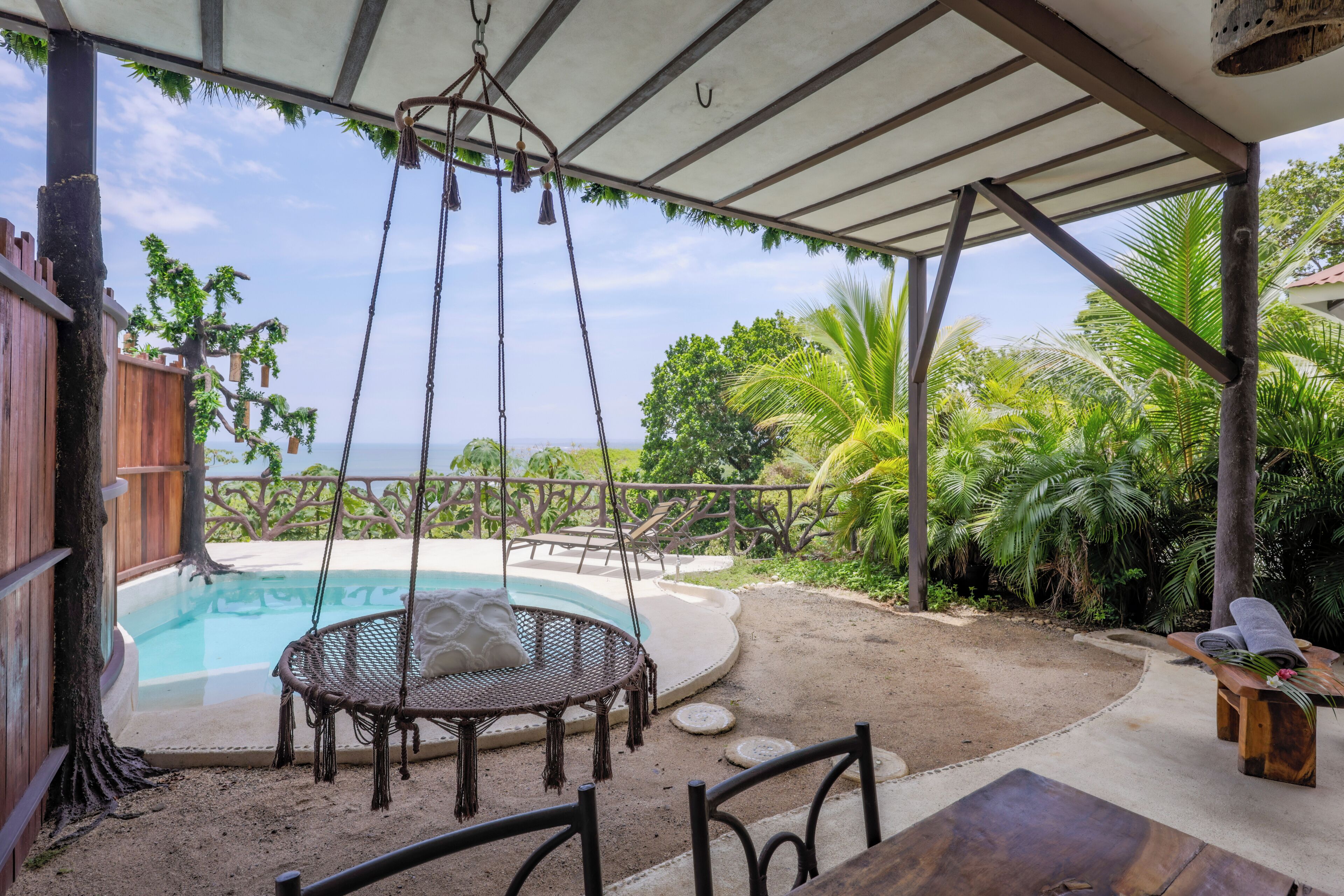 A covered outdoor area with a pool, a swing chair, and a wooden table.