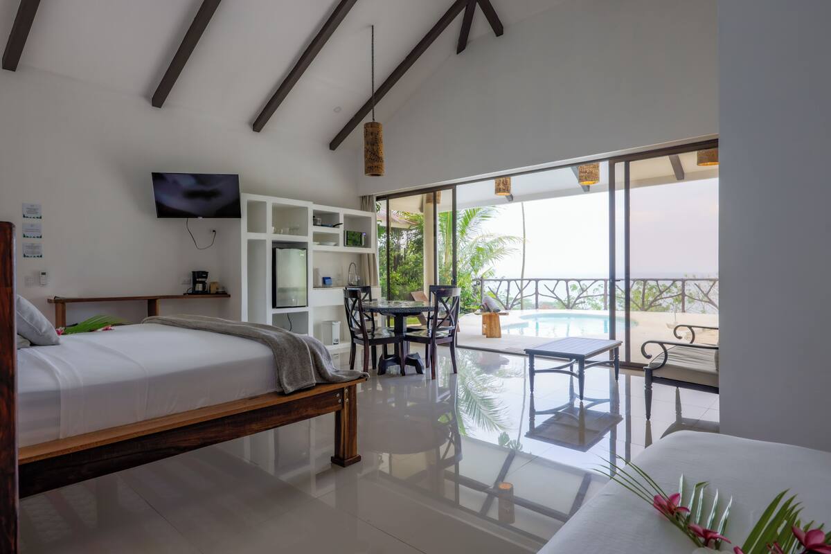 A modern hotel room with a bed, a TV, a dining area with a table and chairs, and a balcony with a pool.