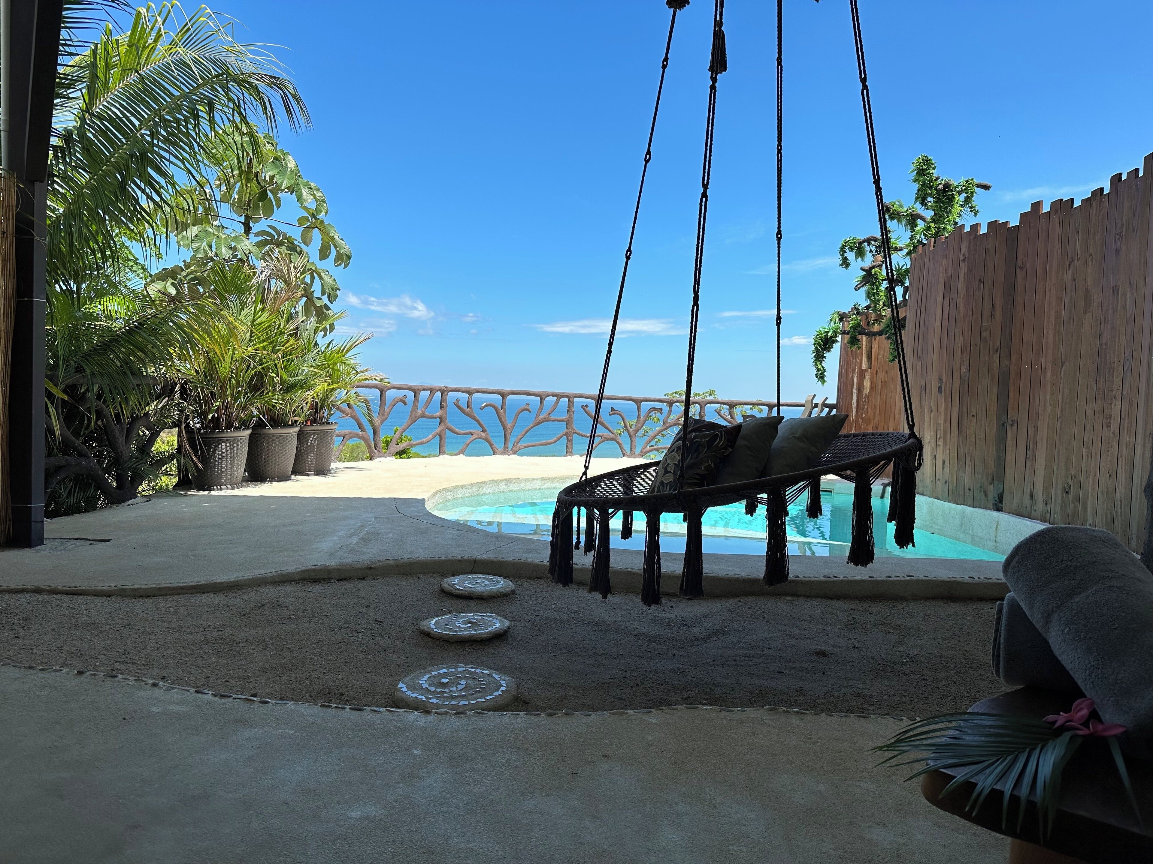 Una silla hamaca junto a una piscina, con un cerco de madera y plantas tropicales.