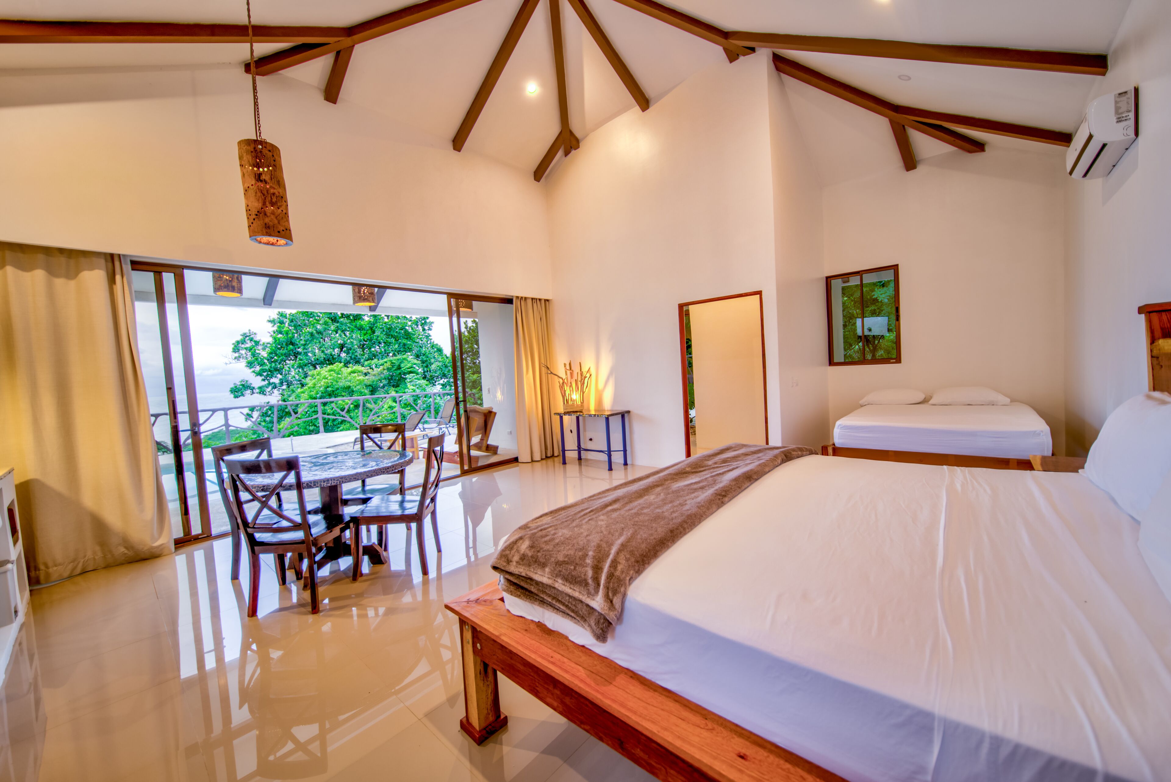 A bedroom with a large bed, a wooden bench, a dining table with chairs, and a view of greenery outside.