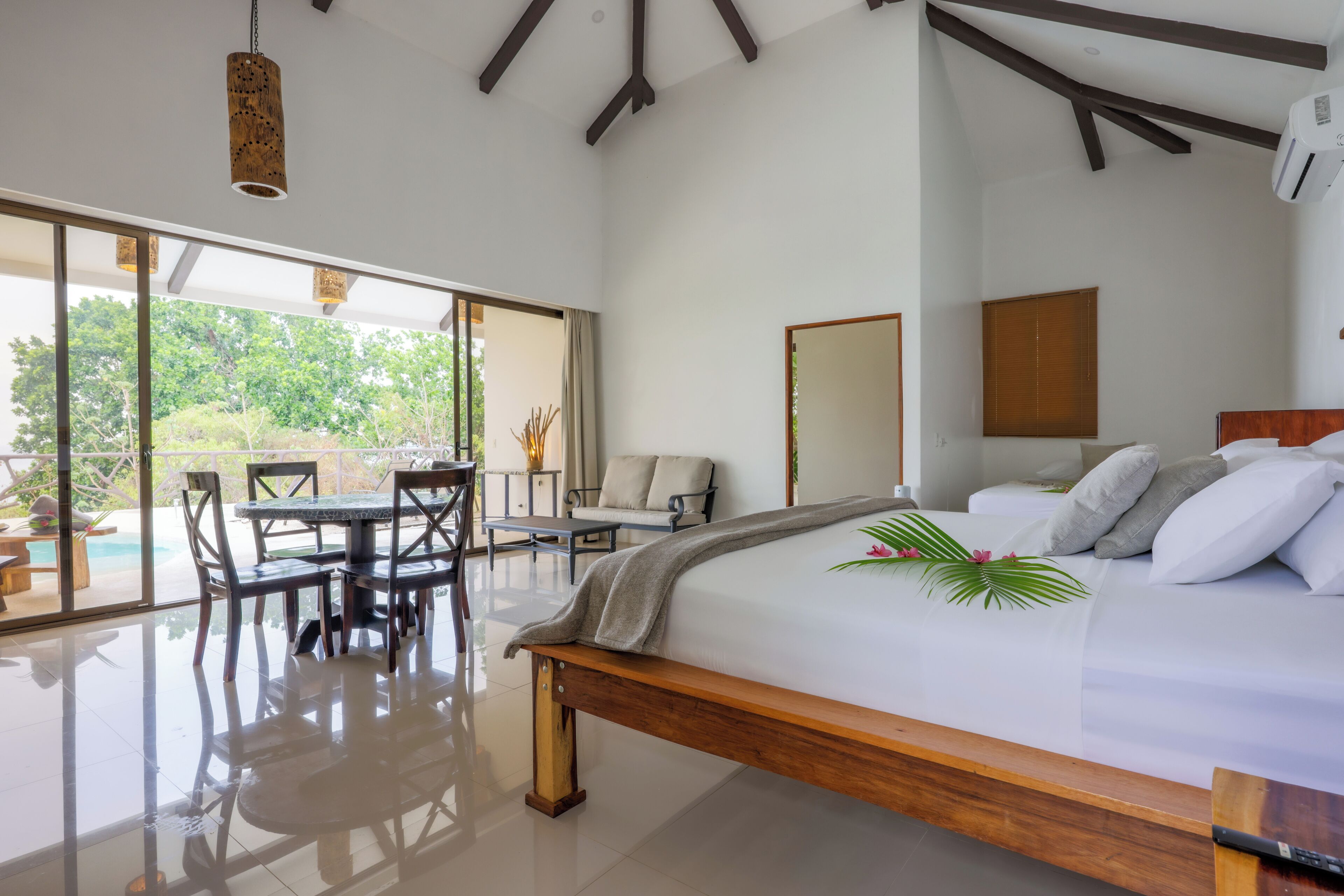 A spacious bedroom with a large bed, a dining area with a table and chairs, and a view of greenery outside.