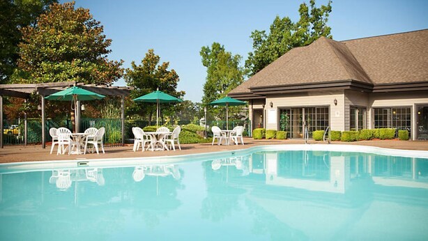 A swimming pool with white lounge chairs and green umbrellas, surrounded by a building with large windows and a covered patio area.