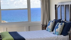 Une chambre avec une grande fenêtre donnant sur la mer, un lit avec une literie bleue et blanche, et un ventilateur de plafond.