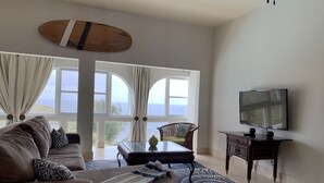 Un salon comprenant un canapé, une table basse et un téléviseur. De grandes fenêtres sont présentes, avec des rideaux, ainsi qu'un ventilateur de plafond. On peut admirer la vue sur l'océan.