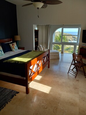 A bedroom with a large bed, a wooden headboard, a ceiling fan, a window with a view of the ocean, a desk, and a chair. - Breathtaking ocean views in Judiths Fancy (Christiansted)