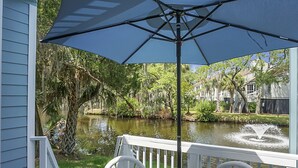 A covered outdoor seating area with a table and chairs, overlooking a calm water body with a fountain, surrounded by lush greenery and trees. - Pet Friendly, 3Bed, easy walk to Pool, Beach, Restaurants, Golf N Pickle Ball  (Edisto Island)