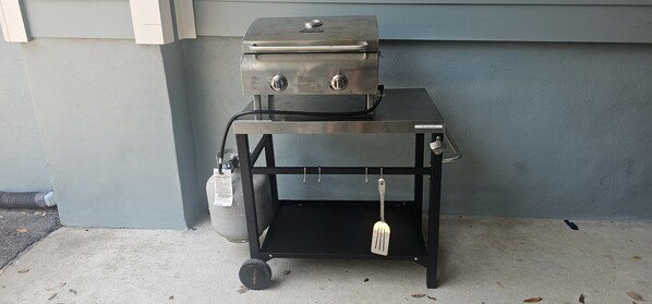 A portable grill on a wheeled cart with a gas tank attached, positioned against a wall.