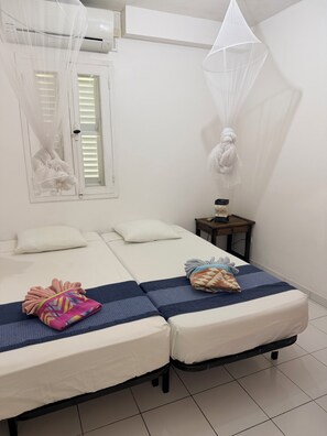 A room with two beds, a small table, a window with blinds, and a hanging mosquito net. - À 10 mn à Pied de la Plage, T3 Jardin vue mer (Les Anses-d'Arlet)
