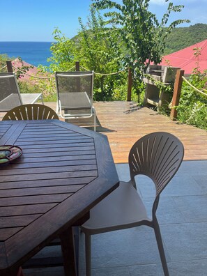 A wooden deck with a table and chairs, overlooking a coastal area with greenery and a red-roofed building. - À 10 mn à Pied de la Plage, T3 Jardin vue mer (Les Anses-d'Arlet)