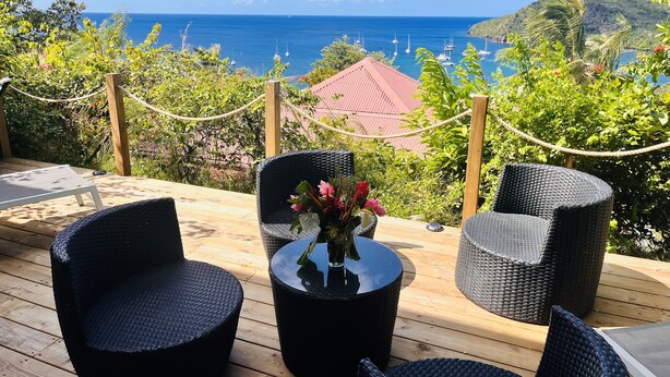 A wooden deck with wicker furniture and a table, overlooking a coastal view with boats and greenery. - À 10 mn à Pied de la Plage, T3 Jardin vue mer (Les Anses-d'Arlet)