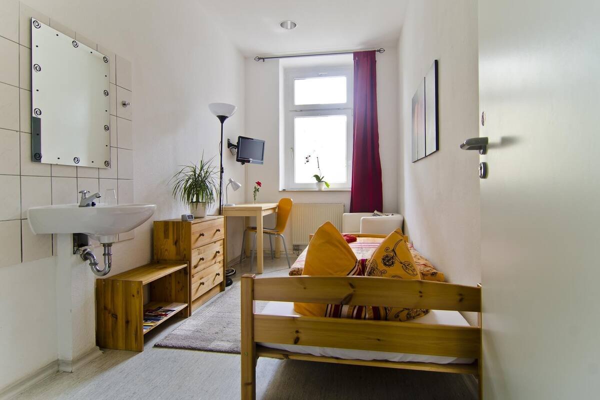 Habitación compacta con cama de madera, escritorio, silla, lavamanos pequeño, espejo y ventana con cortinas.