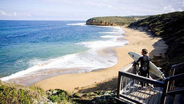 Una playa con una persona cargando una tabla de surf, una escalera que baja a la playa y un litoral rocoso.