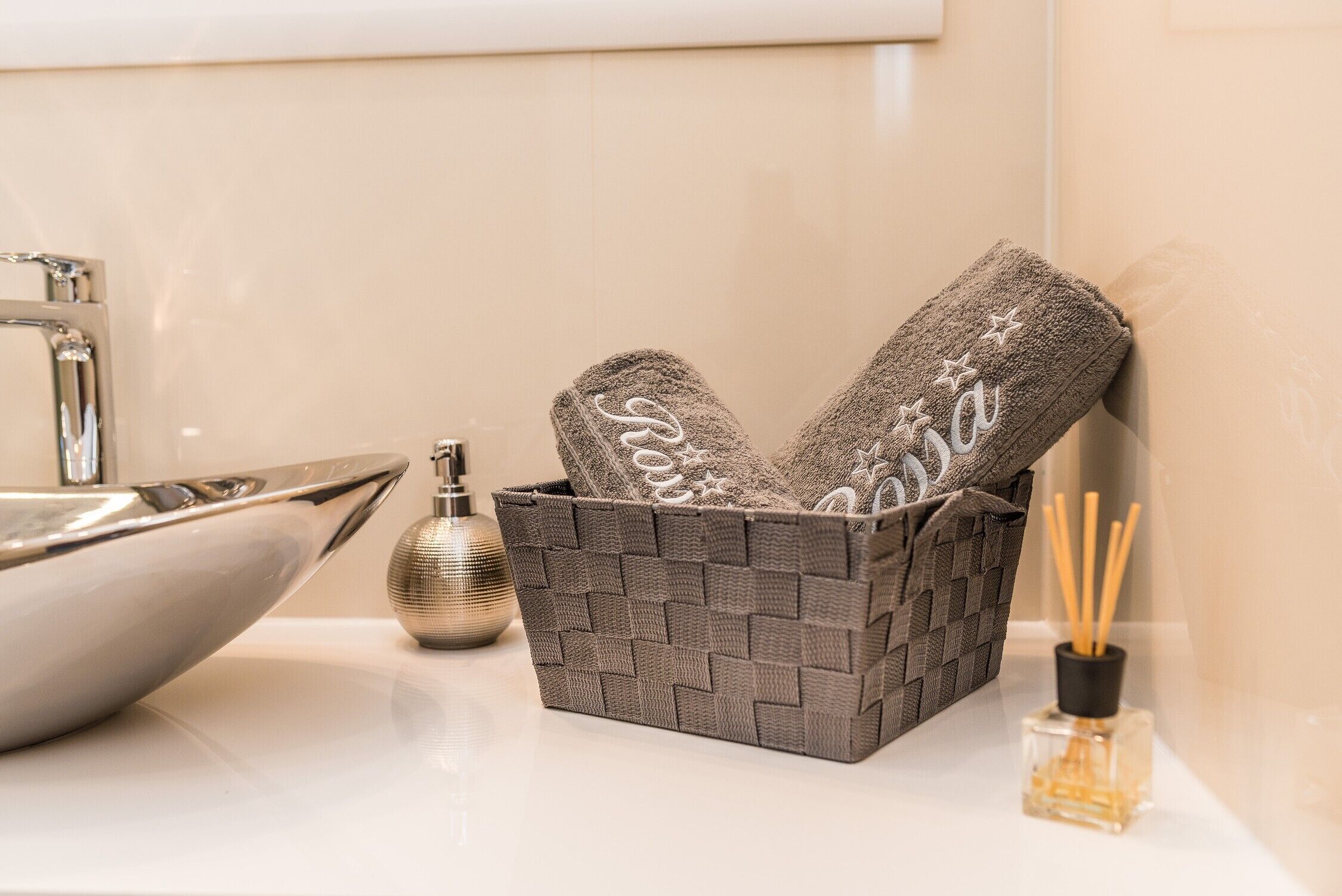 A bathroom with a modern sink, a decorative basket with towels, a bottle of liquid soap, and a reed diffuser.