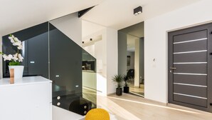 Un intérieur moderne avec un escalier, une console blanche, un pouf jaune et une porte sombre.