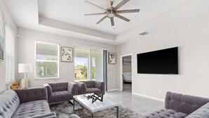 A spacious living room with a ceiling fan, large windows, and a flat-screen TV mounted on the wall. - Luxurious Tampa Bay Area Home in Serene Community! (Riverview)